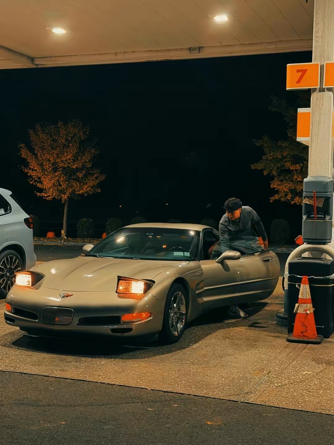 Tan Chevrolet Corvette at gas station at night with attendant refueling, illuminated trees and covered canopy above