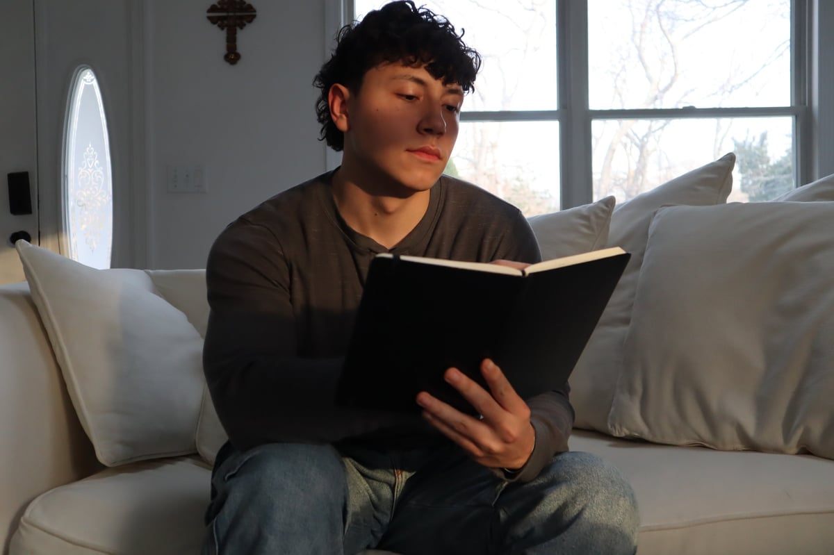 Person with dark curly hair sitting on a couch reading an open book near bright windows
